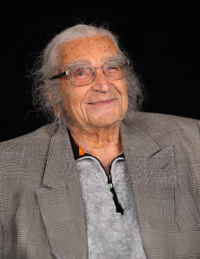 Porträt eines älteren Zeitzeugen vor dunklem Hintergrund, mit Brille, langen grauen Haaren und kariertem Sakko.