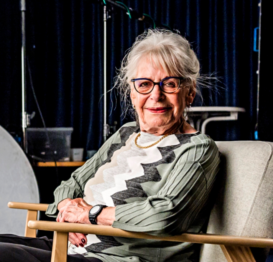 Ältere Zeitzeugin lächelt im Sessel im Studio-Setting, mit grauen Haaren und Brille.