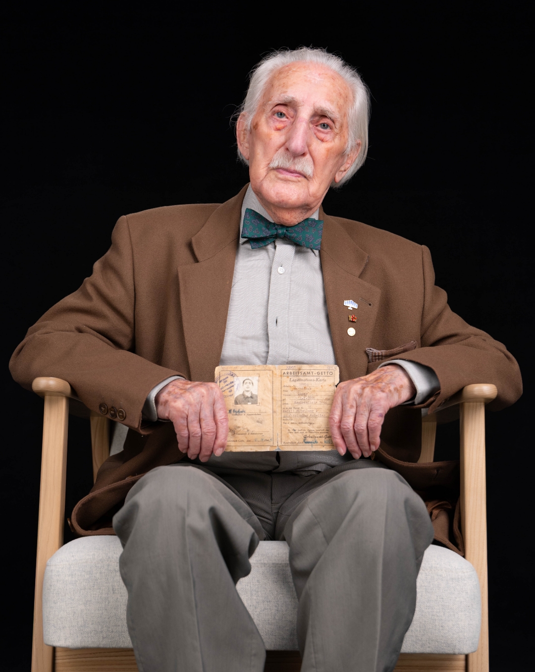 Älterer Zeitzeuge sitzt im Sessel vor schwarzem Hintergrund, mit braunem Sakko und Fliege, Blick nach oben.